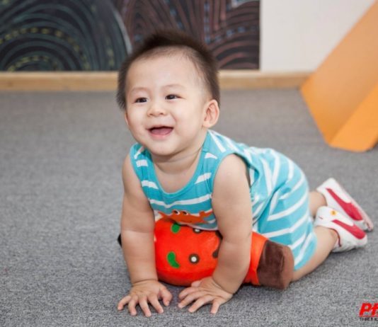 Những Mẫu Thiết Kế Quán Cà Phê Kết Hợp Khu Vui Chơi Trẻ Em. Bé cười đang bò trên thảm xám trong khu vui chơi, mặc bộ liền thân sọc xanh, ôm thú bông cam
