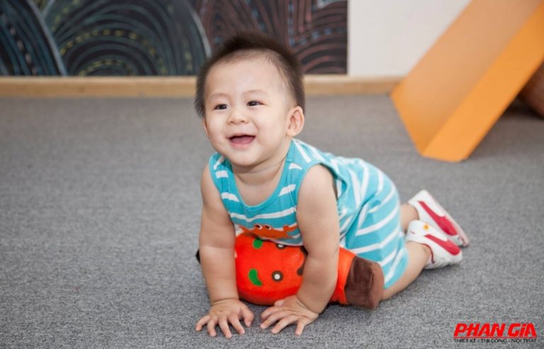 Bé cười đang bò trên thảm xám trong khu vui chơi, mặc bộ liền thân sọc xanh, ôm thú bông cam