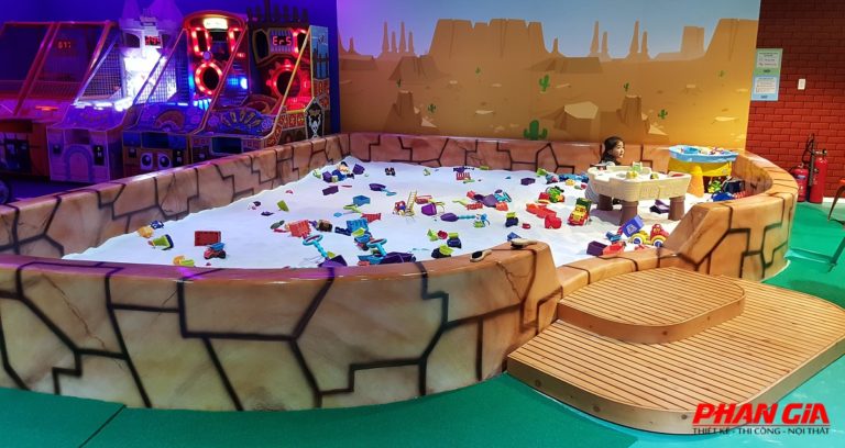 Bể cát trong nhà cho trẻ với cát trắng và đồ chơi nhựa, bé ngồi bên bàn chơi, máy game nền sau