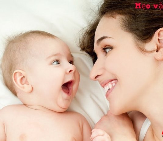 Mẹ mỉm cười nhìn bé sơ sinh đang nằm trên giường, tương tác gần gũi thể hiện chăm sóc trẻ