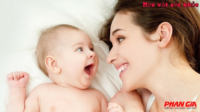 Mẹ mỉm cười nhìn bé sơ sinh đang nằm trên giường, tương tác gần gũi thể hiện chăm sóc trẻ