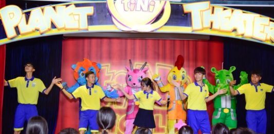 Biểu diễn sân khấu tại tiNiWorld với nhân viên mặc đồng phục vàng-xanh và nhiều mascot màu sắc trước khán giả trẻ em