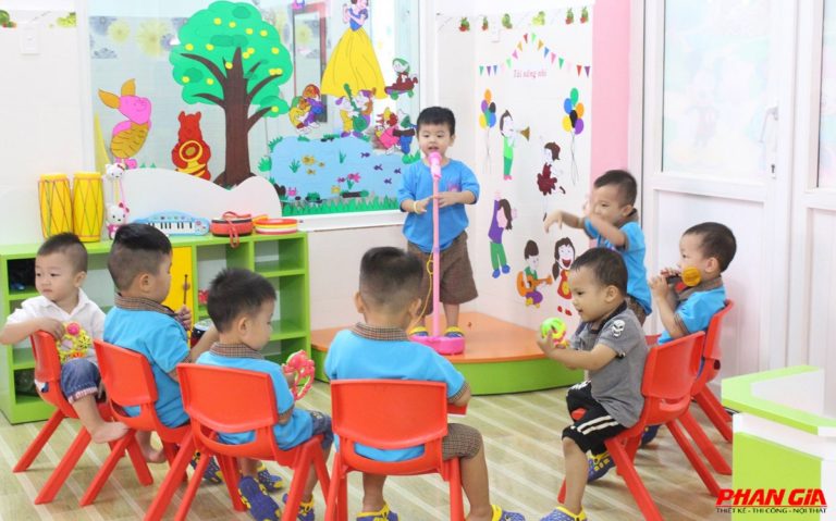Trẻ mầm non ngồi ghế đỏ vòng tròn xem bạn hát trên bục cầm micro đồ chơi