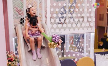 Hai bé gái ôm nhau trượt cầu trượt trong khu vui chơi, một bé cầm bó bong bóng hoa