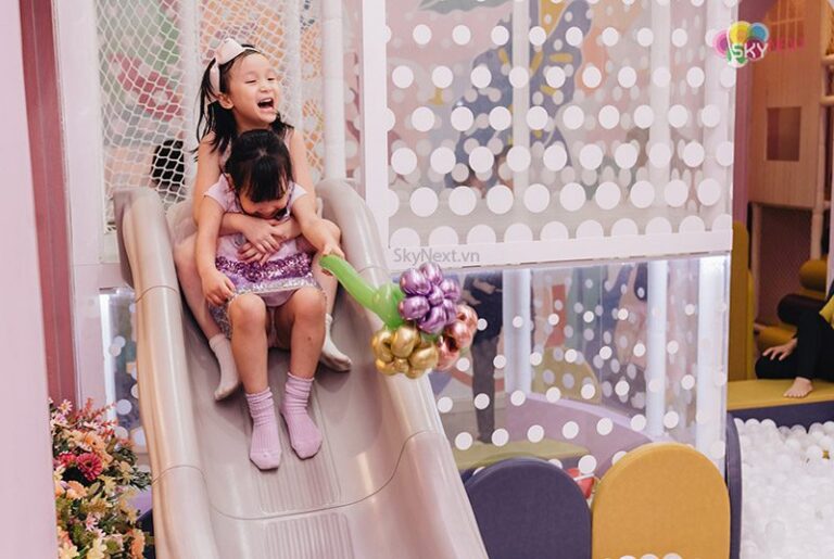Hai bé gái ôm nhau trượt cầu trượt trong khu vui chơi, một bé cầm bó bong bóng hoa