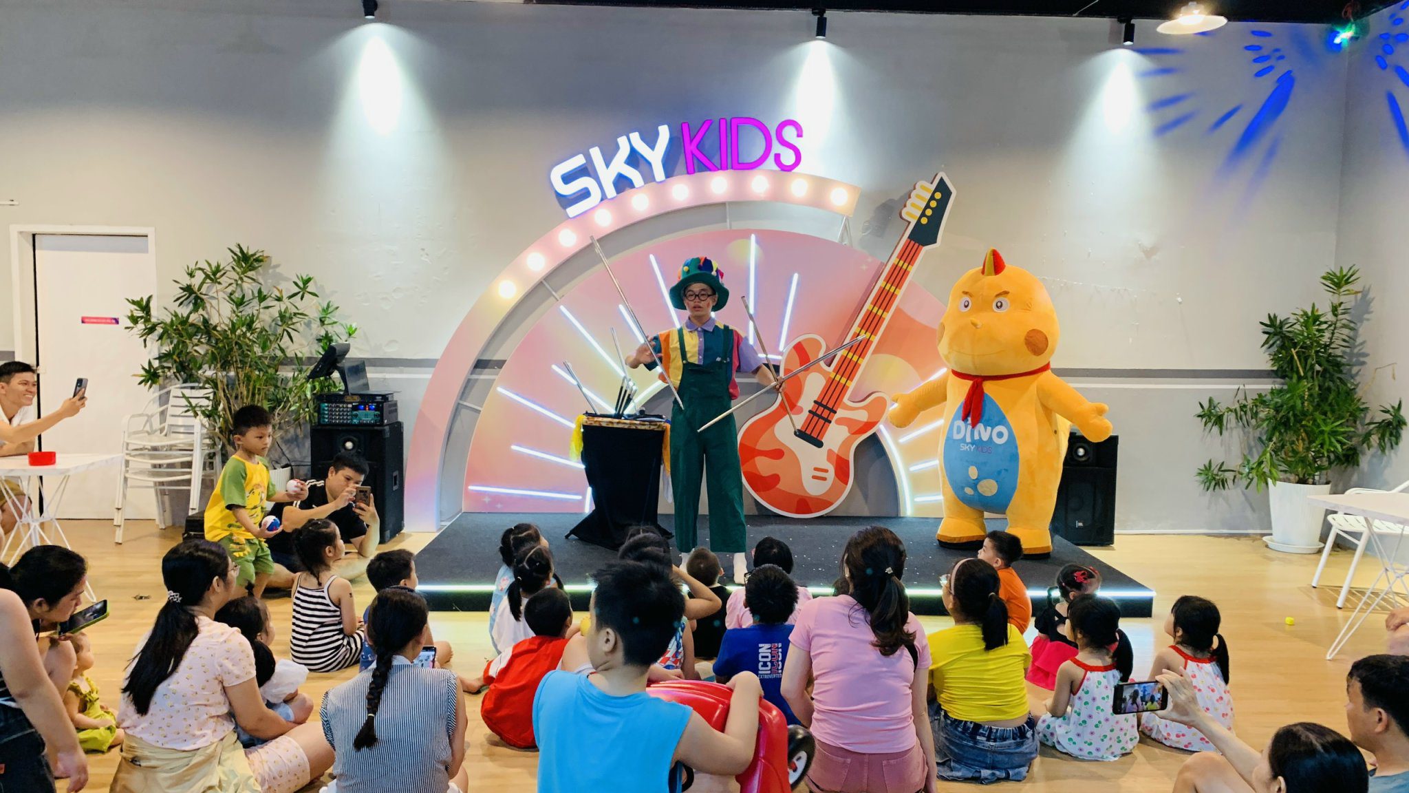 Trẻ em ngồi xem ảo thuật gia trên sân khấu cùng mascot vàng và biển 'SKY KIDS'. họ đang nói về lòng biết ơn